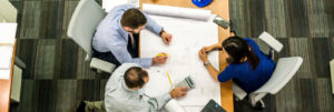 A birds eye view photo of desk with three employees sketching ideas on a large sheet of paper