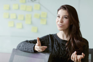 A young female wearing a dark coloured jumper mid conversation with post it notes on a board behind her